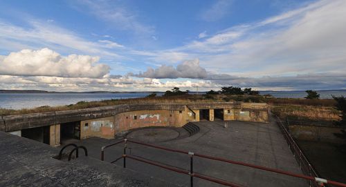 A panoramic view of an abandoned coastal fort with grassy surroundings and a cloudy sky over the water.