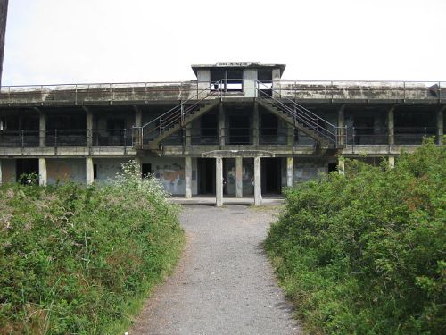 Abandoned building with a central staircase, surrounded by overgrown vegetation and a gravel path leading to the entrance.
