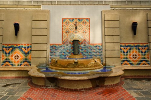 A decorative wall fountain with colorful tiles and a circular stone basin, surrounded by brick flooring.