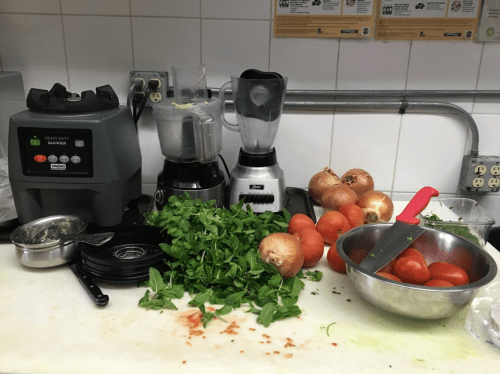 A kitchen counter with blenders, tomatoes, onions, and fresh herbs, ready for meal preparation.