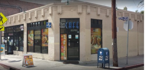 A corner café with large windows displaying colorful menus, located on a city street with nearby signage and a mailbox.