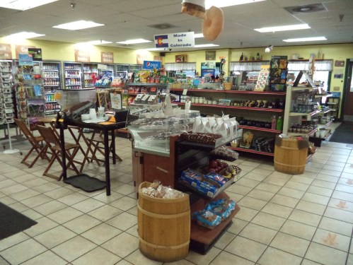 A cozy store interior featuring shelves of snacks, drinks, and local products, with wooden barrels and seating.