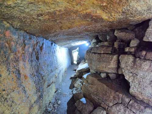 A narrow cave passageway with rough stone walls and a faint light at the end, surrounded by large rock formations.