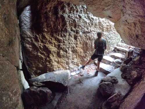 A person stands at the entrance of a cave, looking out into a forested area, with rocky terrain around them.