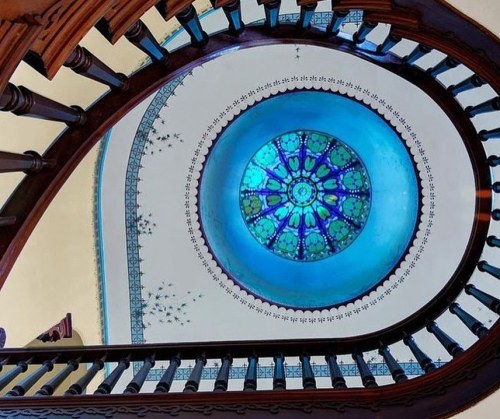 A spiral staircase with wooden railings, leading up to a colorful stained glass dome.
