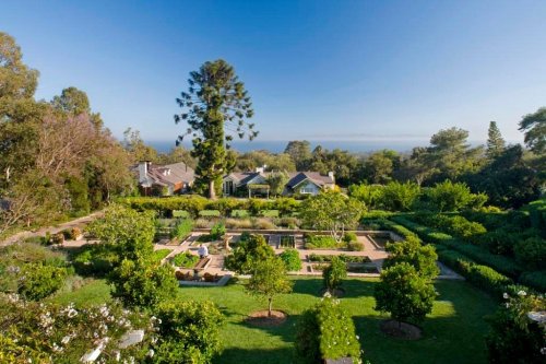 A lush garden with neatly arranged flower beds, trees, and a view of the ocean under a clear blue sky.