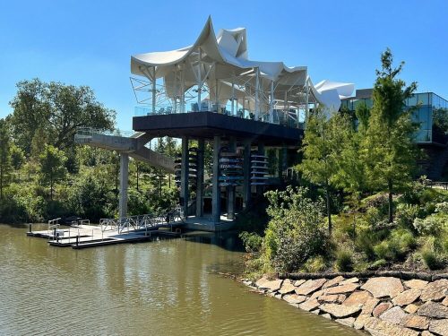 Modern building with a unique roof design, situated by a calm waterway surrounded by greenery.