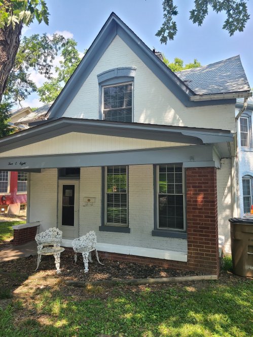 A white, two-story house with a gabled roof, front porch, and two white chairs in the yard. Green trees surround the property.