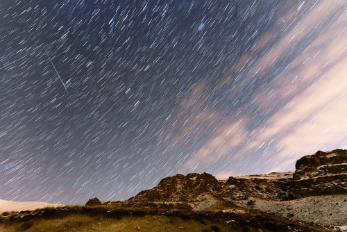 Star trails streak across a night sky above rocky terrain, with clouds softly illuminated by moonlight.