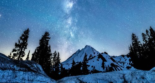 A snowy mountain under a starry night sky, with the Milky Way visible and silhouetted trees in the foreground.