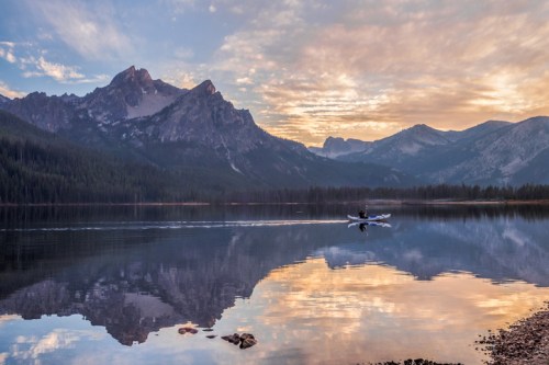 A serene lake at sunrise, with mountains in the background and a person kayaking, reflecting on the water's surface.
