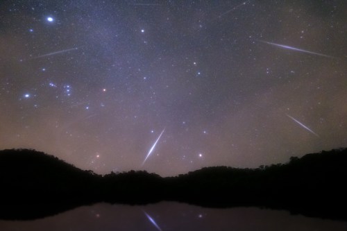 A starry night sky filled with shooting stars above dark silhouettes of mountains and a calm reflective surface.