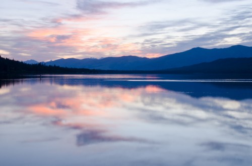 A serene lake at sunset, reflecting colorful clouds and mountains in the background.