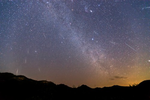 A starry night sky filled with the Milky Way and several shooting stars over dark mountain silhouettes.