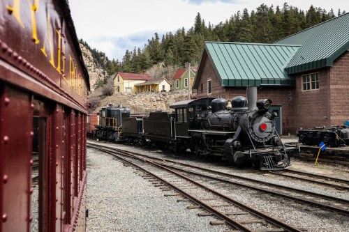 A vintage steam locomotive parked beside a red train car, with a scenic backdrop of trees and buildings.