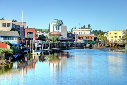 A serene waterfront scene featuring colorful buildings, a wooden pier, and calm water reflecting the clear blue sky.