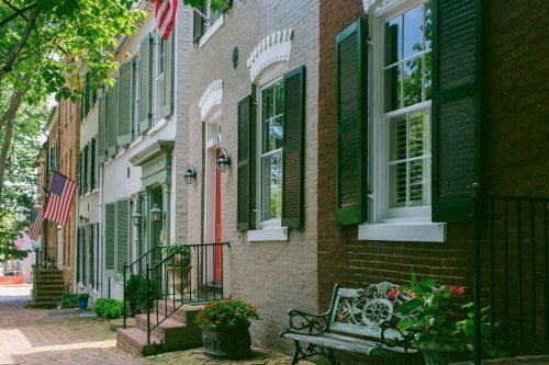 Charming street with historic homes, green shutters, flower pots, and American flags, surrounded by lush trees.