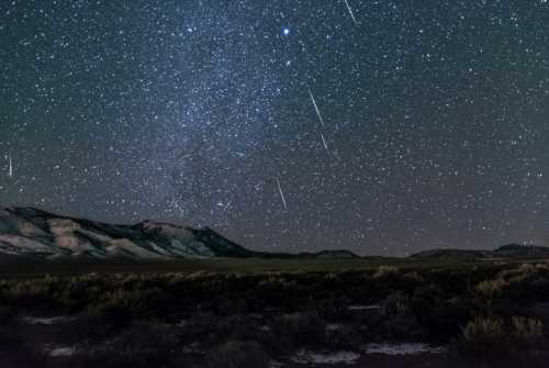 A starry night sky filled with shooting stars over a mountainous landscape and dark foreground.