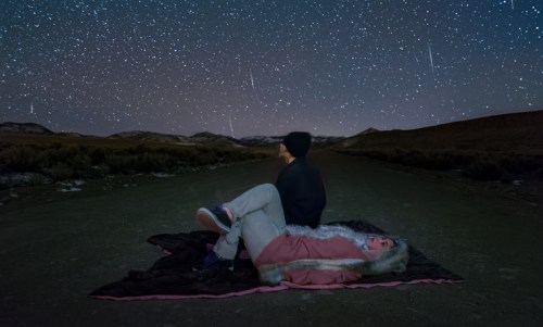 Two people lie on a blanket under a starry sky, watching shooting stars in a remote, dark landscape.