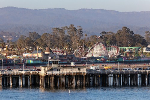 A coastal view of a pier with an amusement park in the background, surrounded by trees and mountains.