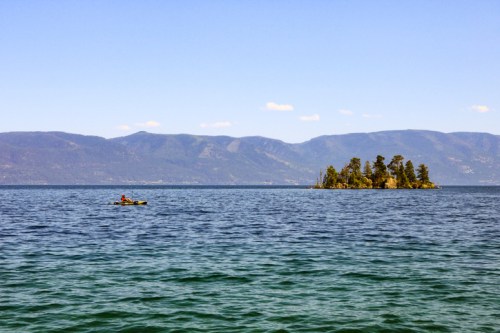 A kayaker paddles on a calm lake near a small island, surrounded by mountains under a clear blue sky.