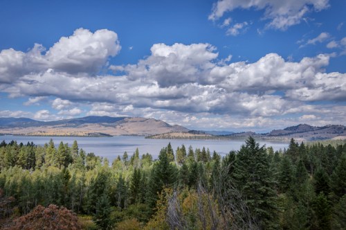 A scenic view of a lake surrounded by mountains and lush green forests under a partly cloudy sky.