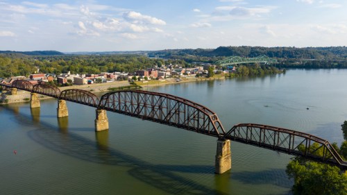 Aerial view of a rusted railway bridge over a river, with a small town and green hills in the background.