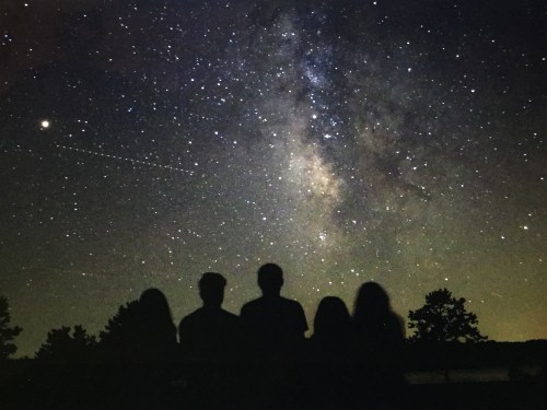 Silhouettes of four people gazing at a starry night sky filled with the Milky Way and shooting stars.