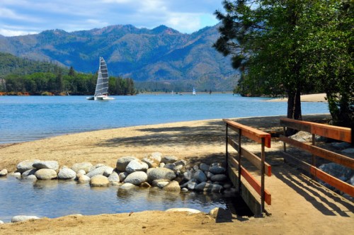 A serene lakeside scene with a wooden bridge, smooth stones, and a sailboat on the water, surrounded by mountains.