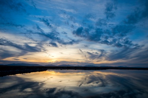 A serene sunset over a calm lake, with vibrant clouds reflecting in the water and mountains in the background.