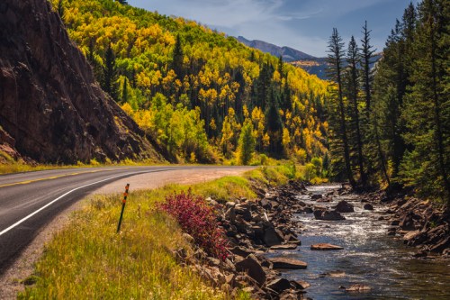 A winding road alongside a river, framed by vibrant autumn foliage and rocky terrain under a clear blue sky.