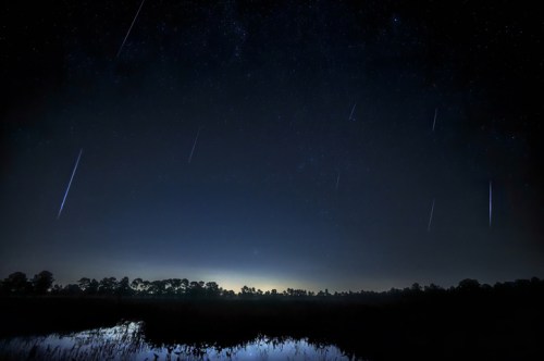 A starry night sky with multiple shooting stars over a dark landscape and calm water reflecting the scene.