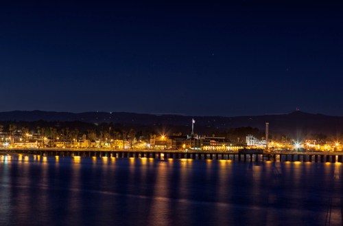 Nighttime view of a waterfront city with illuminated buildings and reflections on the water. Mountains in the background.