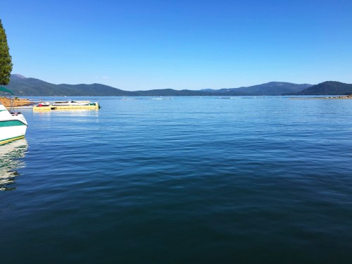 A calm lake with clear blue water, surrounded by mountains under a bright blue sky, with boats docked nearby.
