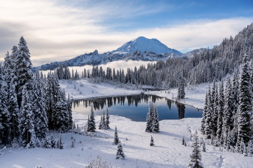 A serene winter landscape featuring a snow-covered mountain, evergreen trees, and a reflective lake under a blue sky.