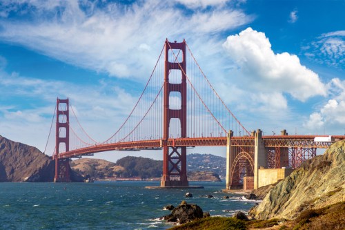 Golden Gate Bridge spanning a bay, with blue skies and clouds in the background, surrounded by hills and water.