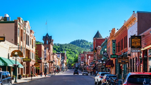A vibrant street scene with historic buildings, shops, and signs, set against a backdrop of green hills and a clear blue sky.