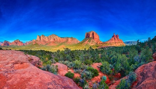 A panoramic view of red rock formations and lush greenery under a bright blue sky.
