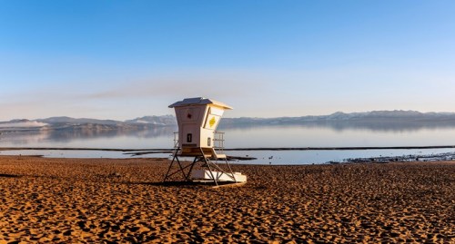 A lifeguard tower on a sandy beach with calm water and mountains in the background under a clear blue sky.