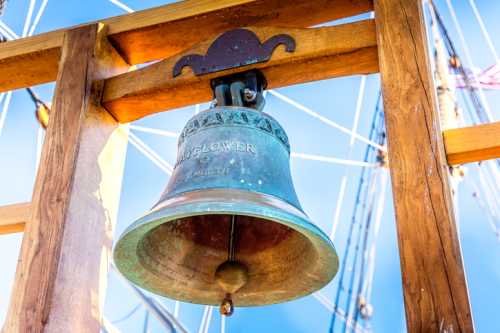 Close-up of a ship's bell inscribed "Mayflower," hanging from a wooden structure against a blue sky.