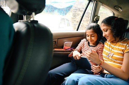 Two girls sitting in a car, sharing a tablet and smiling, with rain visible on the window.