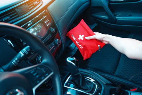 A hand reaches for a red first aid kit inside a car's interior, near the gear shift and dashboard.