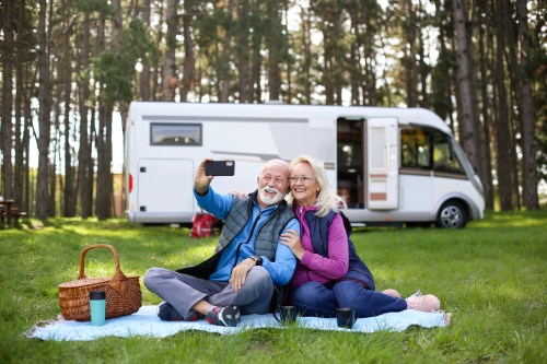 An elderly couple sits on a picnic blanket, smiling and taking a selfie in front of a camper in a forested area.
