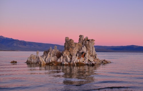 A rocky formation rises from calm water, with a pastel sunset sky and distant mountains in the background.