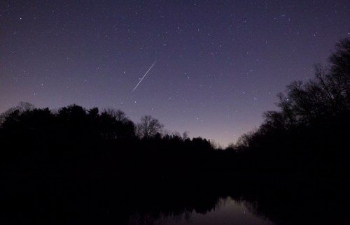 A serene night sky filled with stars, reflecting on a calm lake, with a meteor streaking across the scene.