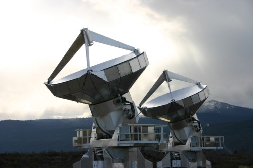 Two large satellite dishes point towards the sky, set against a cloudy backdrop with distant mountains.
