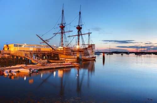 A historic ship docked at a marina during sunset, reflecting in calm waters with boats nearby.