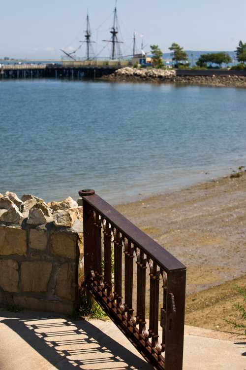 A rusted gate leads to a sandy shore with a calm water view and a tall ship docked in the background.