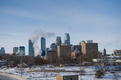 A winter cityscape featuring tall buildings, smoke rising, and a snowy foreground under a clear blue sky.
