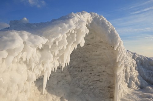 Icicles hang from a frozen arch, with a clear blue sky in the background, showcasing a winter landscape.
